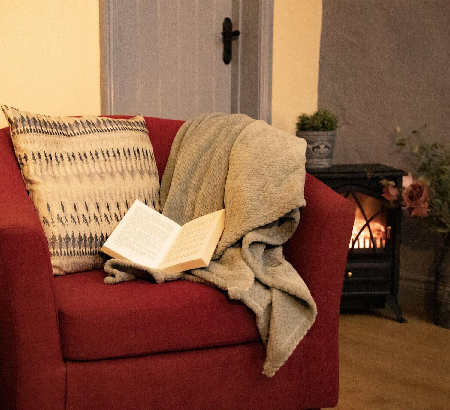 An open book on a red chair with a blanket draped over the arm and a lit fireplace in the background