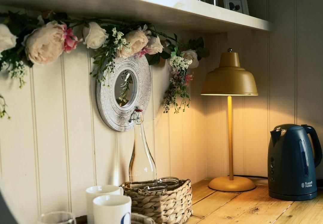 flower-decorated interior with a kettle, mugs, a glass bottle of water, and a lit lamp